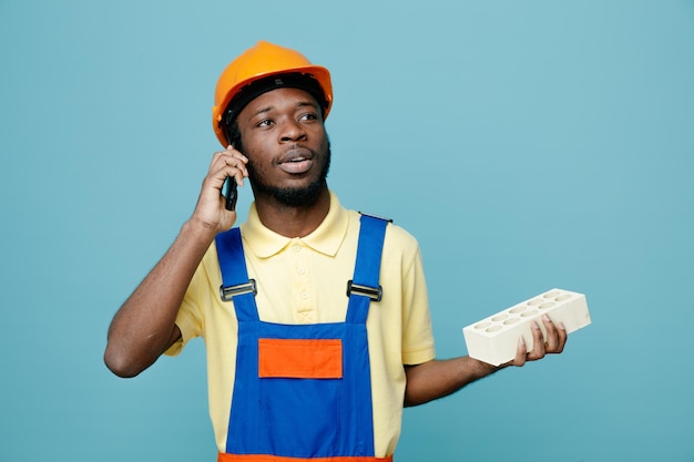 Pensando en sostener ladrillo joven constructor afroamericano en uniforme habla por teléfono aislado sobre fondo azul.