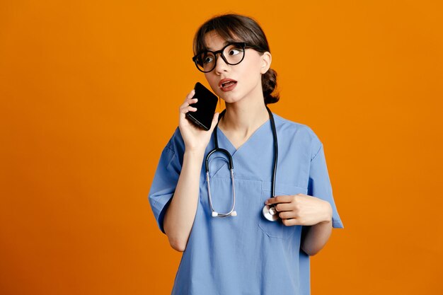 Pensando en sostener el teléfono joven doctora vistiendo uniforme fith estetoscopio aislado sobre fondo naranja