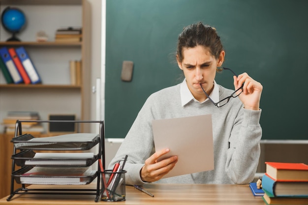 pensando sosteniendo gafas y mirando papel en su mano joven maestro sentado en el escritorio con herramientas escolares en el aula