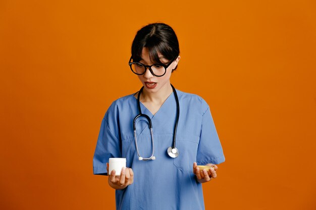 Pensando sosteniendo lata de píldora joven doctora vistiendo uniforme fith estetoscopio aislado sobre fondo naranja