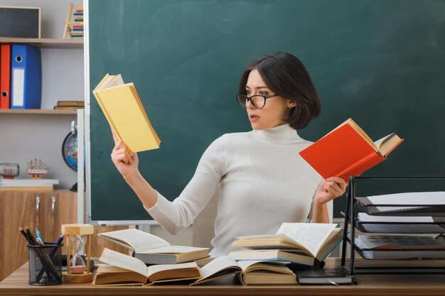 pensando sosteniendo y mirando el libro joven maestra sentada en el escritorio con herramientas escolares en el aula