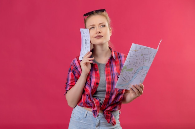 Pensando viajero joven vestida con camisa roja y gafas en la cabeza sosteniendo el mapa con boleto sobre fondo rosa aislado