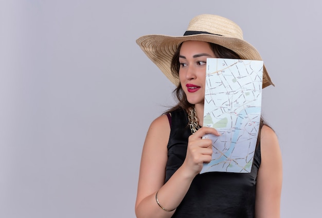 Pensando viajero joven vestida con camiseta negra con sombrero sosteniendo mapa sobre fondo blanco.