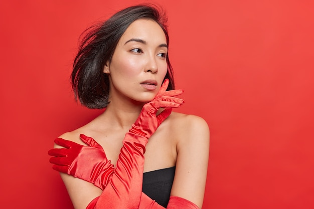 Pensativa hermosa dama asiática usa vestido negro, guantes largos, vestidos para ocasiones especiales, se ve pensativamente lejos, tiene el cabello oscuro flotando en el aire aislado sobre una pared roja vívida