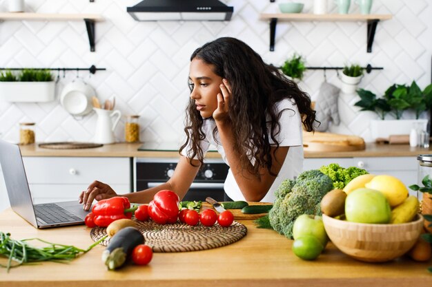 Pensativa hermosa mujer mulata está mirando en la pantalla del portátil en la cocina moderna en la mesa llena de verduras y frutas, vestida con una camiseta blanca