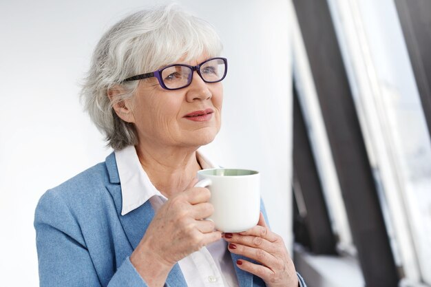 Pensativa hermosa pensionista con elegantes anteojos rectangulares y chaqueta azul con taza, disfrutando del aroma de un buen capuchino fresco. Mujer senior elegante pelo gris bebiendo té