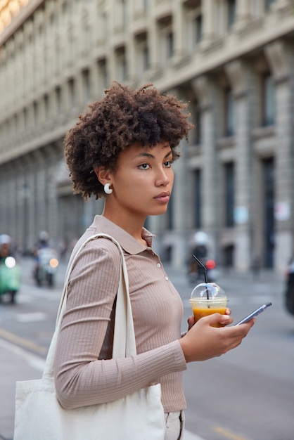 Pensativa joven mujer de pelo rizado sostiene un teléfono inteligente pasea por la ciudad envía mensajes de texto, bebidas, charlas de cócteles en línea, vestida de manera informal, lleva una bolsa de tela, se encuentra en el centro de la ciudad disfruta del tiempo libre