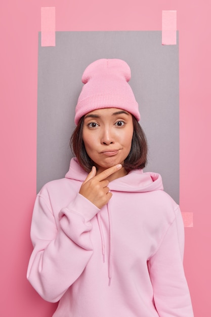 Pensativa mujer asiática milenaria sostiene la barbilla tiene expresión dudosa, considera una sugerencia interesante vestida con una sudadera con capucha informal y poses de sombrero contra la pared rosa con una hoja de papel enlucida