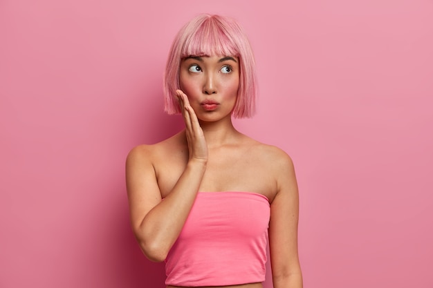 Pensativa mujer de cabello rosado toca la mejilla, mira hacia arriba, concentrada en algo, vestida con una camiseta sin mangas informal, muestra los hombros desnudos, tiene el cabello rosado a la moda