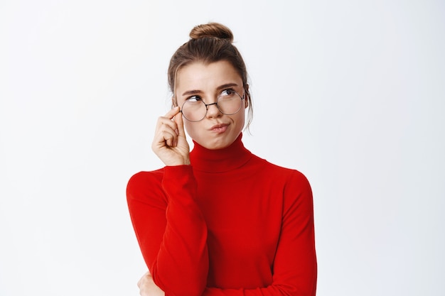 Pensativa mujer guapa con gafas pensando, mirando hacia el lado derecho en el espacio vacío y reflexionando, de pie pensativo contra la pared blanca