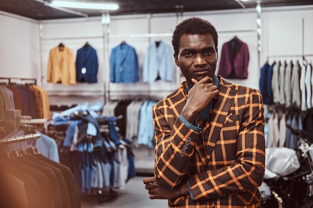 Foto gratuita pensativo hombre afroamericano elegantemente vestido posando con la mano en la barbilla mientras está de pie en una tienda clásica de ropa de hombre, mirando a la cámara.