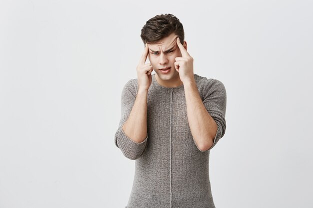 Pensativo hombre guapo caucásico vestido con suéter, mantiene los dedos en las sienes, mira seriamente, reflexiona, trata de encontrar la decisión adecuada en una situación difícil. Gente, juventud, concepto de estilo de vida