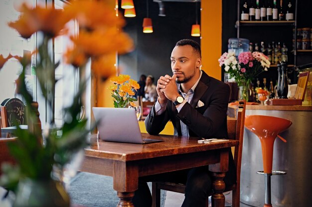 Un pensativo hombre negro estadounidense vestido con un traje elegante usando una computadora portátil en un café.