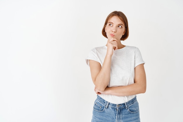 Pensativo idea de maquillaje de mujer joven, mirando a un lado con mirada reflexiva, tocando la barbilla, pensando mientras está de pie en la pared blanca