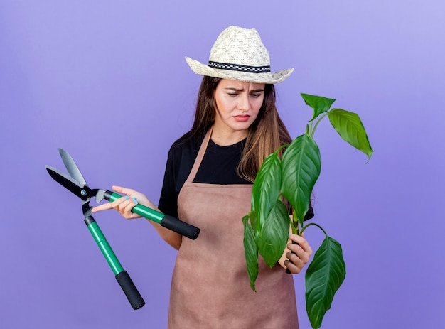 Pensativo jardinero hermosa chica en uniforme con sombrero de jardinería sosteniendo tijeras y mirando la planta en su mano aislado en azul