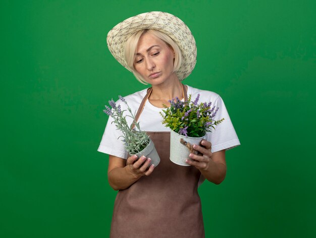 Pensativo jardinero rubio de mediana edad mujer en uniforme con sombrero sosteniendo y mirando macetas aisladas en la pared verde con espacio de copia
