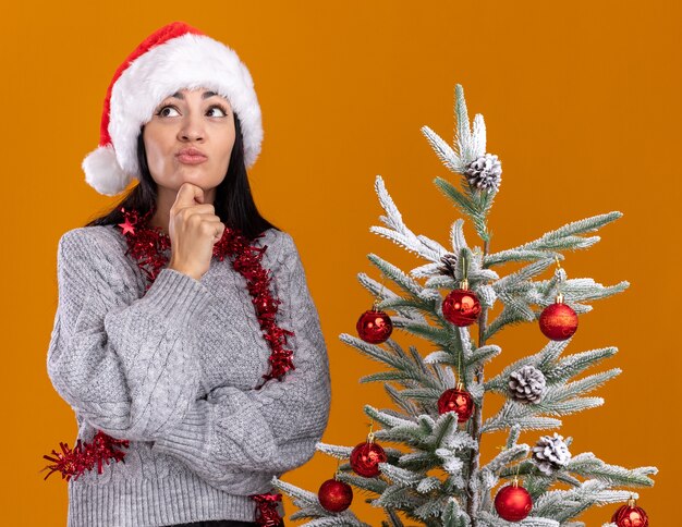 Pensativo joven caucásica vistiendo gorro de navidad y guirnalda de oropel alrededor del cuello de pie cerca del árbol de navidad decorado tocando la barbilla mirando hacia arriba aislado sobre fondo naranja