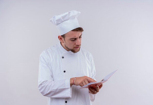 Un pensativo joven chef barbudo hombre vestido con uniforme de cocina blanco y sombrero mirando portátil en una pared blanca