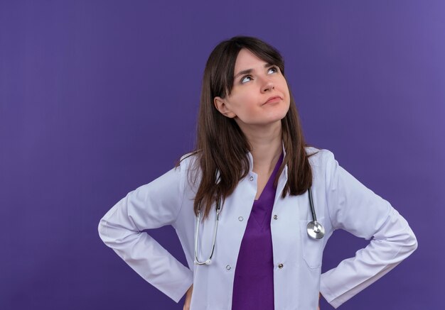 Pensativo joven doctora en bata médica con estetoscopio pone las manos en la cintura y mira hacia arriba sobre fondo violeta aislado con espacio de copia