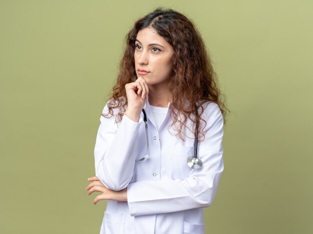 Pensativo joven doctora vistiendo bata médica y estetoscopio manteniendo la mano en la barbilla mirando al lado aislado en la pared verde oliva con espacio de copia