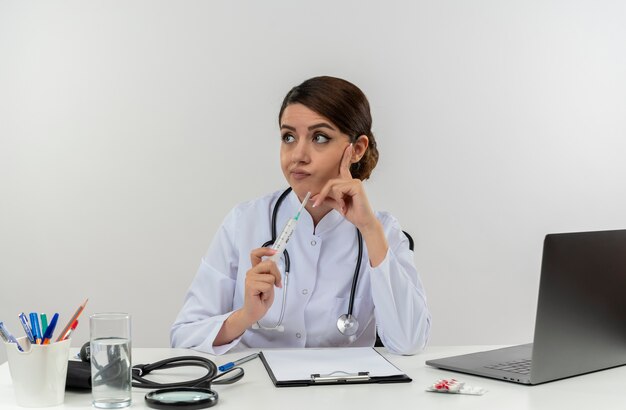 Pensativo joven doctora vistiendo bata médica y estetoscopio sentado en el escritorio con herramientas médicas y portátil sosteniendo una jeringa mirando de lado manteniendo el dedo en la cara aislada