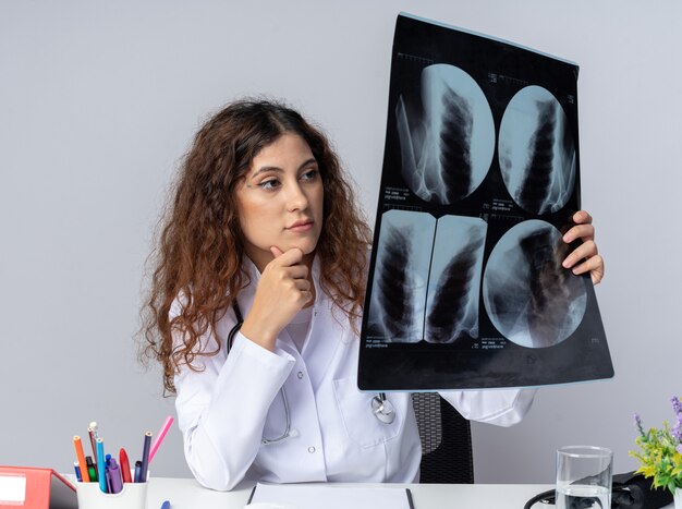 Pensativo joven doctora vistiendo bata médica y estetoscopio sentado en la mesa con herramientas médicas manteniendo la mano en la barbilla sosteniendo y mirando la toma de rayos X aislada en la pared blanca