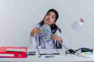 Foto gratuita pensativo joven doctora vistiendo bata médica y un estetoscopio sentados en el escritorio con herramientas médicas sosteniendo dinero mirando al lado aislado