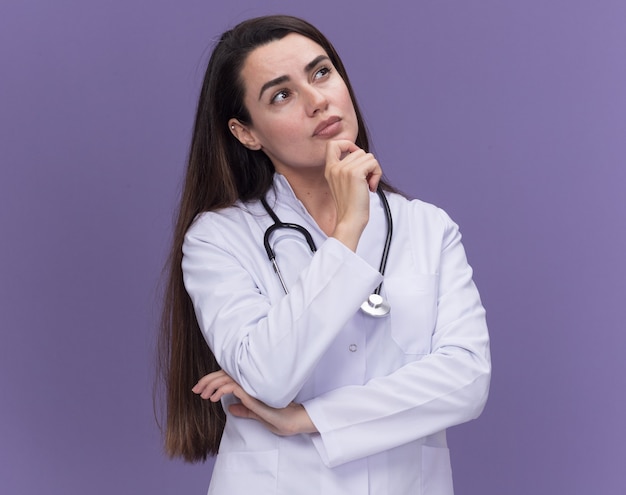 Pensativo joven doctora vistiendo bata médica con estetoscopio sostiene la barbilla mirando hacia arriba