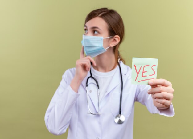 Pensativo joven doctora vistiendo bata médica, máscara y estetoscopio y estirando la nota de sí poniendo el dedo en la mejilla en la pared verde aislada