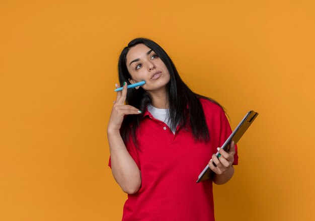 Pensativo joven morena caucásica vestida con camisa roja tiene lápiz y portapapeles aislado en la pared naranja