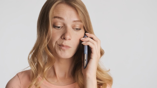 Pensativo joven mujer rubia hablando por teléfono inteligente sobre fondo blanco.