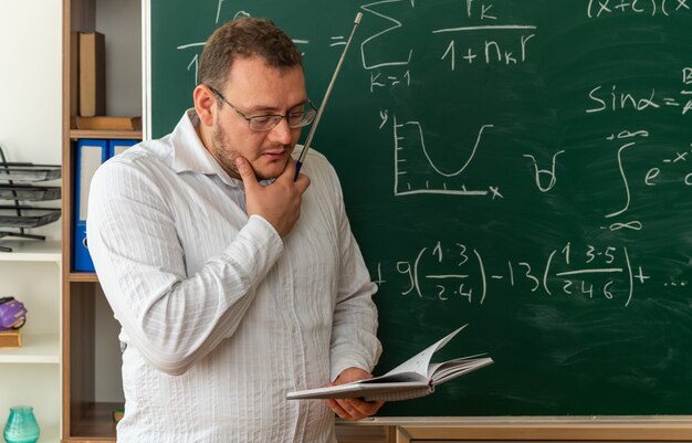 Pensativo joven profesor con gafas de pie delante de la pizarra en el aula manteniendo la mano en la barbilla sosteniendo el puntero leyendo el bloc de notas