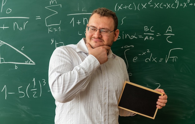 Pensativo joven profesor con gafas de pie delante de la pizarra en el aula sosteniendo una mini pizarra manteniendo la mano en la barbilla mirando al lado