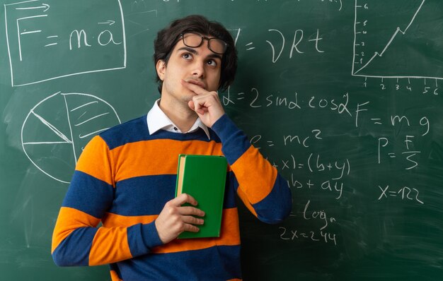 Pensativo joven profesor de geometría con gafas en la frente de pie delante de la pizarra en el aula sosteniendo el libro cerrado tocando el labio mirando hacia arriba