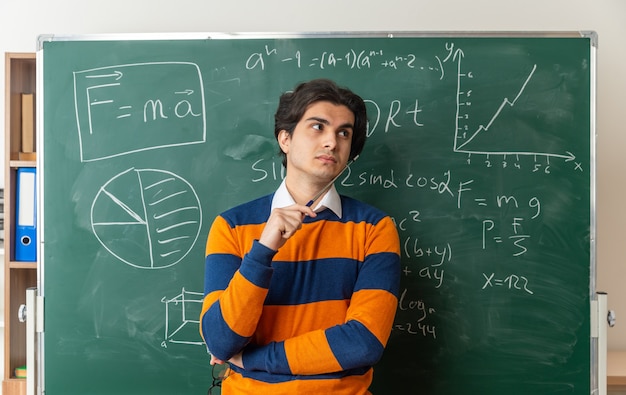 Pensativo joven profesor de geometría de pie delante de la pizarra en el aula mirando al lado sosteniendo el palillo del puntero