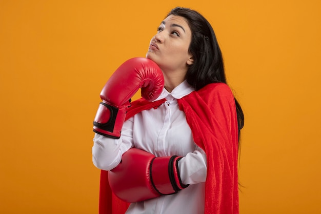 Pensativo joven superhéroe caucásica chica con guantes de caja poniendo la mano en la barbilla mirando hacia arriba aislado sobre fondo naranja con espacio de copia