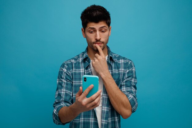 Pensativo joven tocando el labio sosteniendo y mirando el teléfono móvil aislado sobre fondo azul.