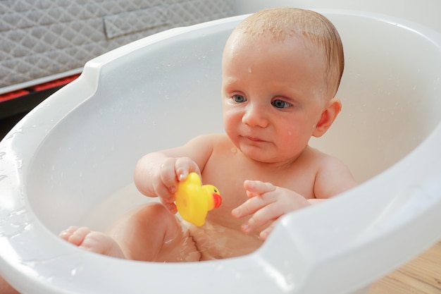 Pensativo lindo bebé mojado sosteniendo y jugando pato de juguete de goma amarillo mientras tiene bañera en casa. Fotografía de cerca. Concepto de cuidado infantil o salud