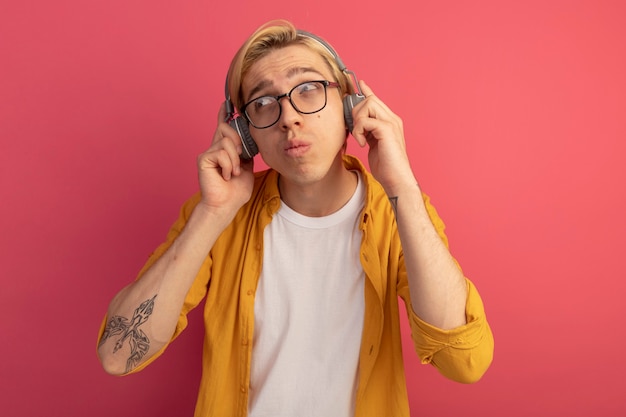 Pensativo mirando al lado joven chico rubio vistiendo camiseta amarilla y gafas con auriculares aislados en rosa