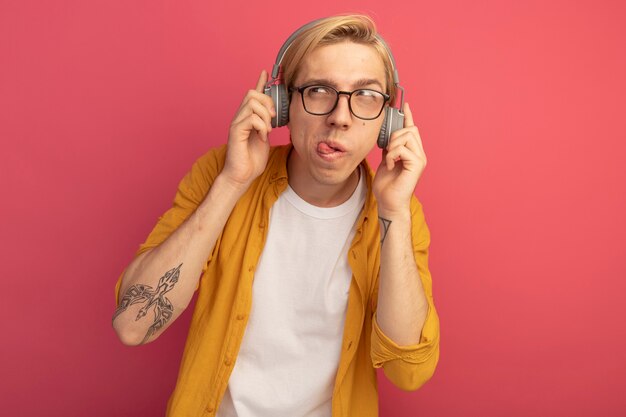 Pensativo mirando al lado joven chico rubio vistiendo camiseta amarilla y gafas con auriculares mostrando la lengua aislada en rosa con espacio de copia