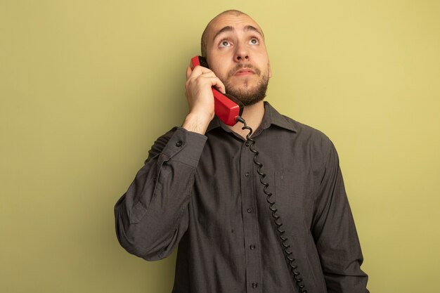 Pensativo mirando hacia arriba joven guapo vistiendo camiseta negra habla por teléfono aislado en verde oliva
