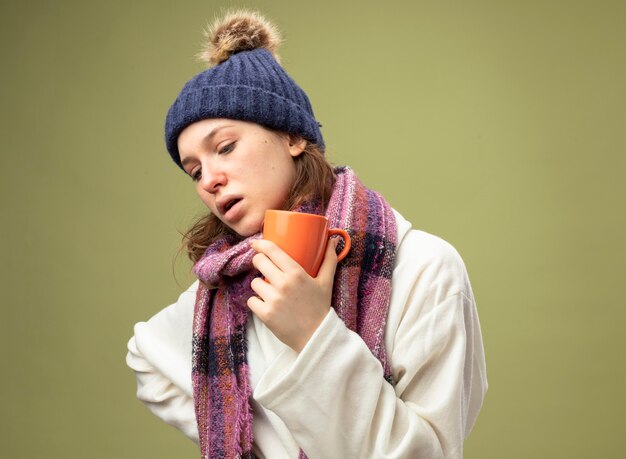 Pensativo niña enferma mirando downwearing bata blanca y gorro de invierno con bufanda sosteniendo una taza de té aislado en verde oliva