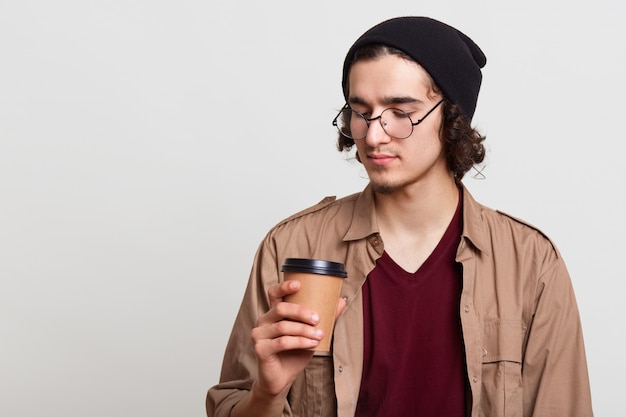 Pensativo pensativo yougster tomando una taza de café, sosteniendo una bebida caliente en una mano, mirándolo atentamente, posando aislado en gris claro, estando en descanso. Concepto de juventud.