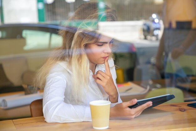 Pensativo profesional femenino sentado en el escritorio en el espacio de trabajo conjunto o cafetería, usando tableta