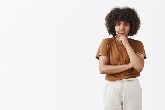 Pensativo, sospechoso y dudoso linda adolescente afroamericana con peinado afro en camiseta marrón sosteniendo la mano en la barbilla frunciendo el ceño mientras piensa mirando con incredulidad y sonrisa