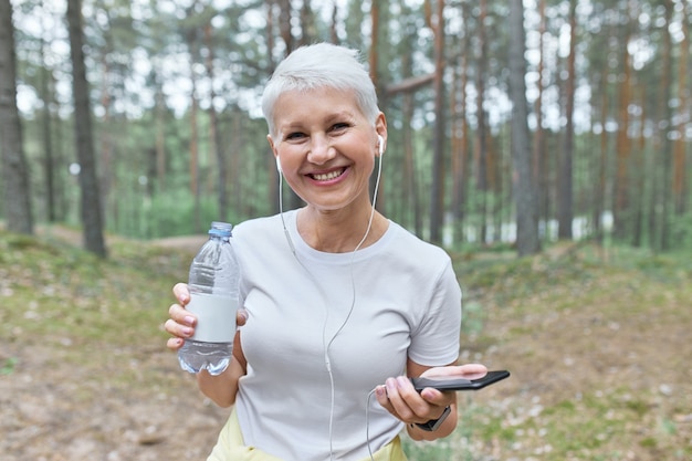 Pensionista alegre que descansa después de un entrenamiento cardiovascular al aire libre, posando en un bosque de pinos con el móvil y una botella de agua, refrescándose, escuchando música