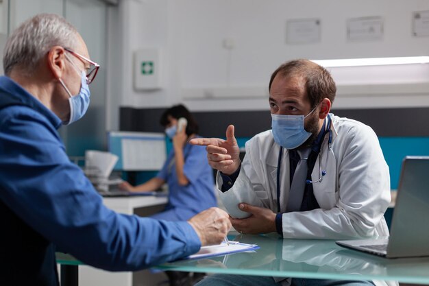 Pensionista firma informe de examen en gabinete médico con médico, con mascarilla. Anciano haciendo la firma en el formulario, reuniéndose con el médico de cabecera en la consulta de control.