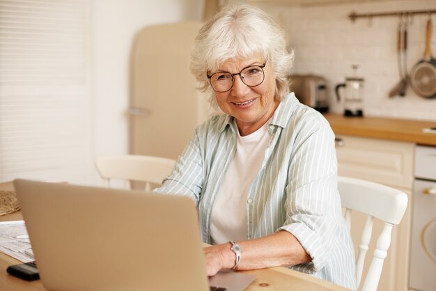 Pensionista mujer autónoma seria atractiva que trabaja lejos de casa, sentado en la cocina frente a la computadora portátil abierta, usando anteojos. Concepto de personas, edad, trabajo y ocupación