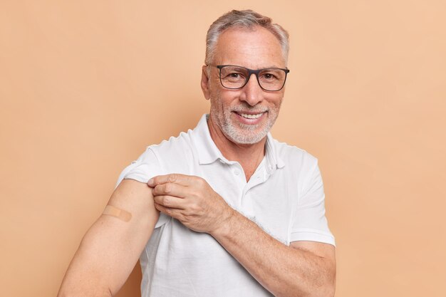 Un pensionista de pelo gris muestra que el brazo enyesado se vacunó para reducir el riesgo de contraer o propagar el coronavirus, espera efectos secundarios, usa anteojos transparentes y poses de camiseta en el interior de la clínica.