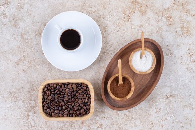 Una pequeña bandeja con tazones de azúcar y café molido en polvo junto a una taza de café y un tazón de granos de café.